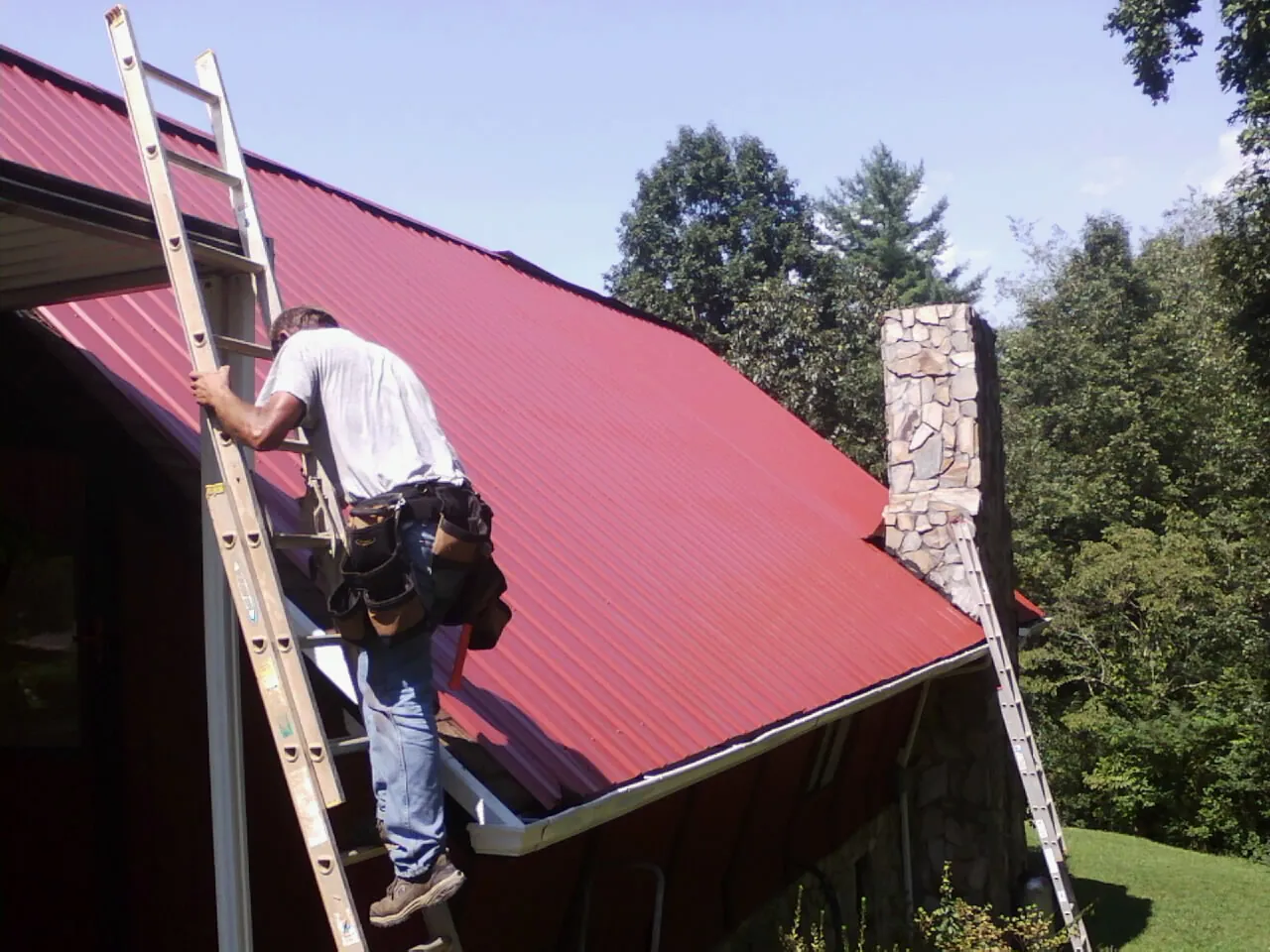 Roofer on a ladder beside a red metal roof and stone chimney — Soffit & Fascia Repair by Roofing Brothers in Frostburg